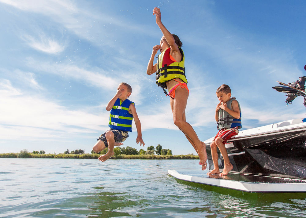 3 young kids in life vets jumping off the back side of a boat into a cool, blue lake