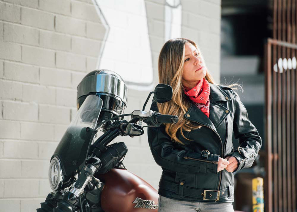 Woman with long brown hair wearing a black leather jacket standing next to her motorcycle. Helmet is on the handlebars.